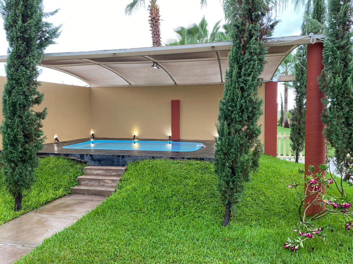 A shaded area features a small plunge pool, surrounded by vibrant green grass. Palm and other trees provide additional greenery, while soft lighting enhances the space. A stone pathway leads to the entrance of the area, creating a serene outdoor setting.