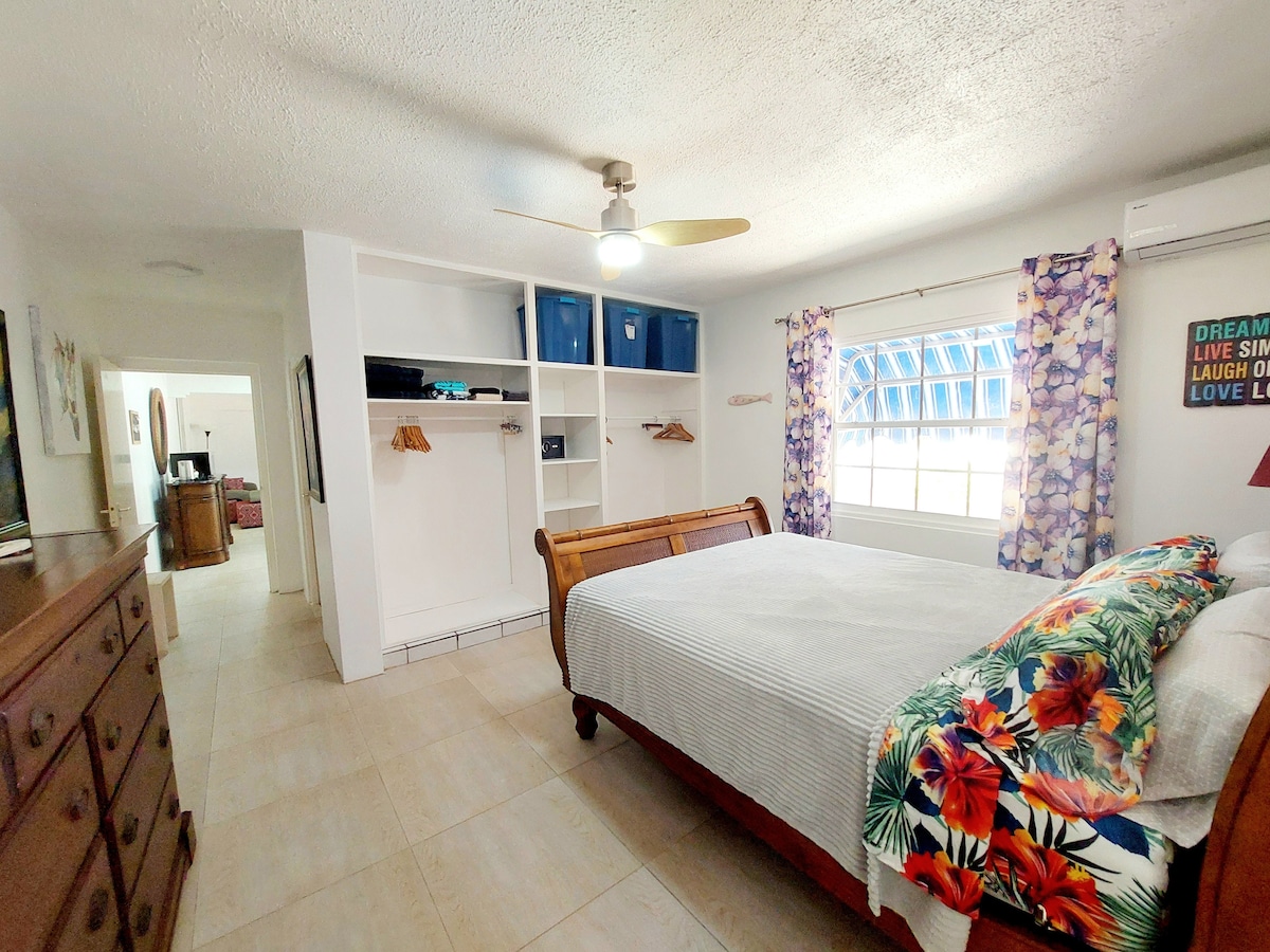 A spacious bedroom is furnished with a queen bed, adorned with a colorful floral throw blanket. Shelves are present for storage, with a ceiling fan providing overhead cooling. Natural light filters through a large window, complemented by vibrant purple curtains.