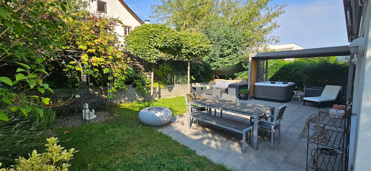 A serene garden area features a spacious wooden table with accompanying chairs, surrounded by lush greenery. A hot tub sits under a shaded pergola, while a lounging area is positioned nearby. Sunlight bathes the stone patio, highlighting the tranquil atmosphere.