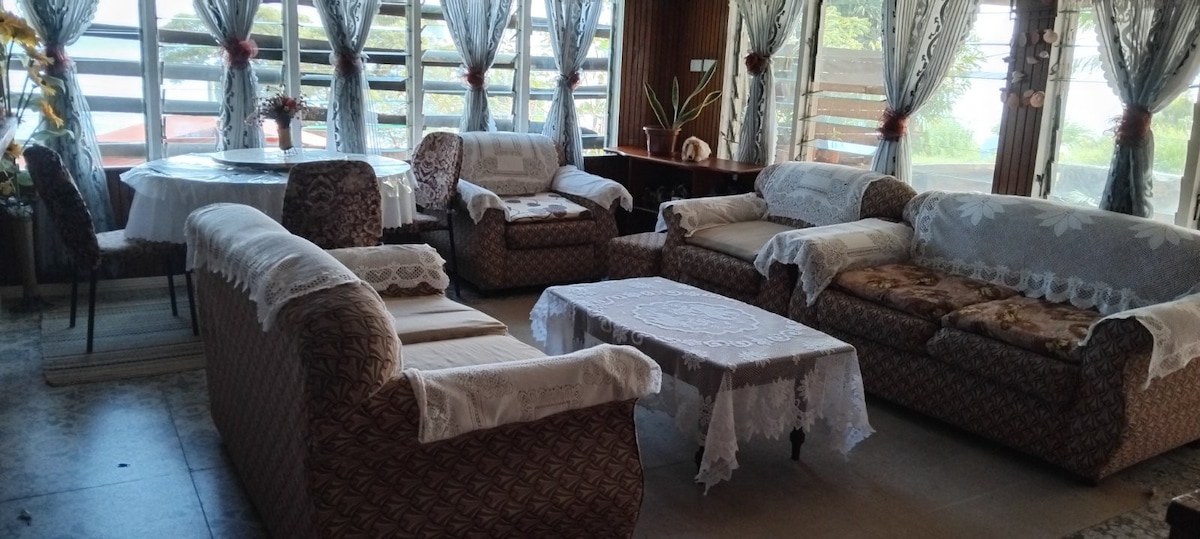 A comfortable living area is arranged with patterned sofas and armchairs featuring decorative coverings. A glass-top coffee table is situated in the center, surrounded by plants. Large windows with sheer curtains allow natural light to flow in, offering views of the outdoor scenery.