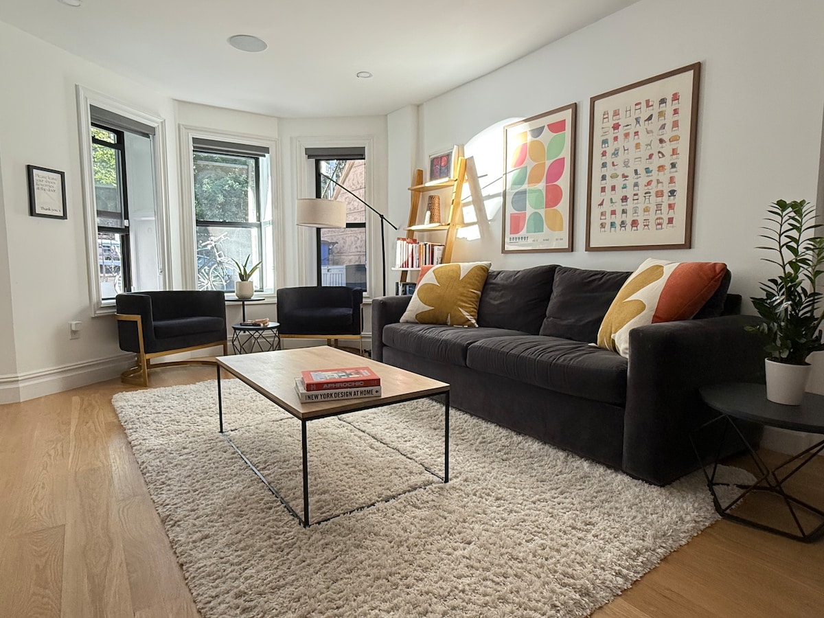 A spacious living area features a dark sectional sofa adorned with colorful throw pillows. A coffee table sits centrally on a soft area rug. Floor-to-ceiling windows provide natural light, while a plant adds a touch of greenery. Artwork decorates the walls, enhancing the design.