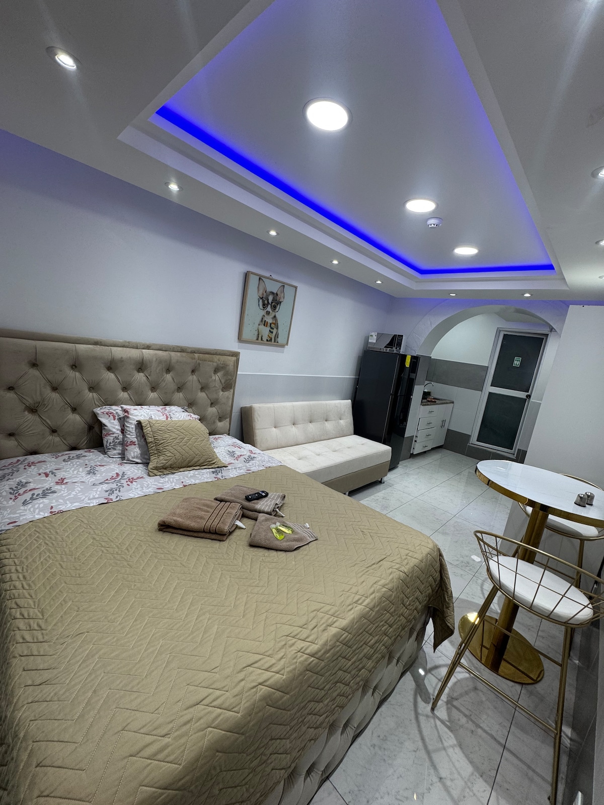 A well-organized studio space is showcased, featuring a spacious bed with neutral bedding and a decorative headboard. A white sofa sits adjacent to the bed. Modern appliances, including a refrigerator, are visible, along with a small dining table and chairs under warm lighting and a blue-accented ceiling.