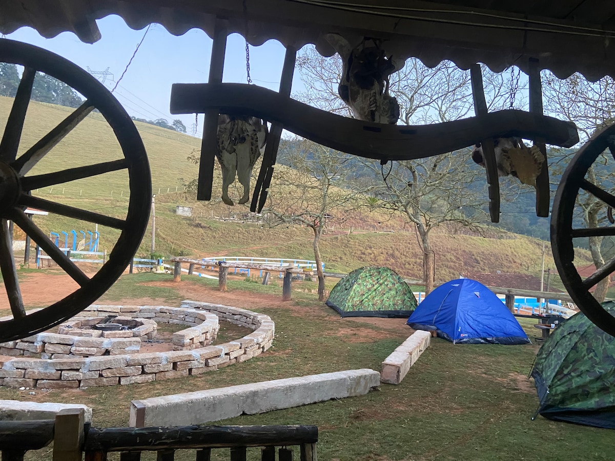 The view from a covered porch includes hanging wooden decorations and a scenic landscape. In the foreground, stone seating creates a circular arrangement. Two blue and green tents can be seen pitched on the grassy area, with rolling hills and trees in the background.