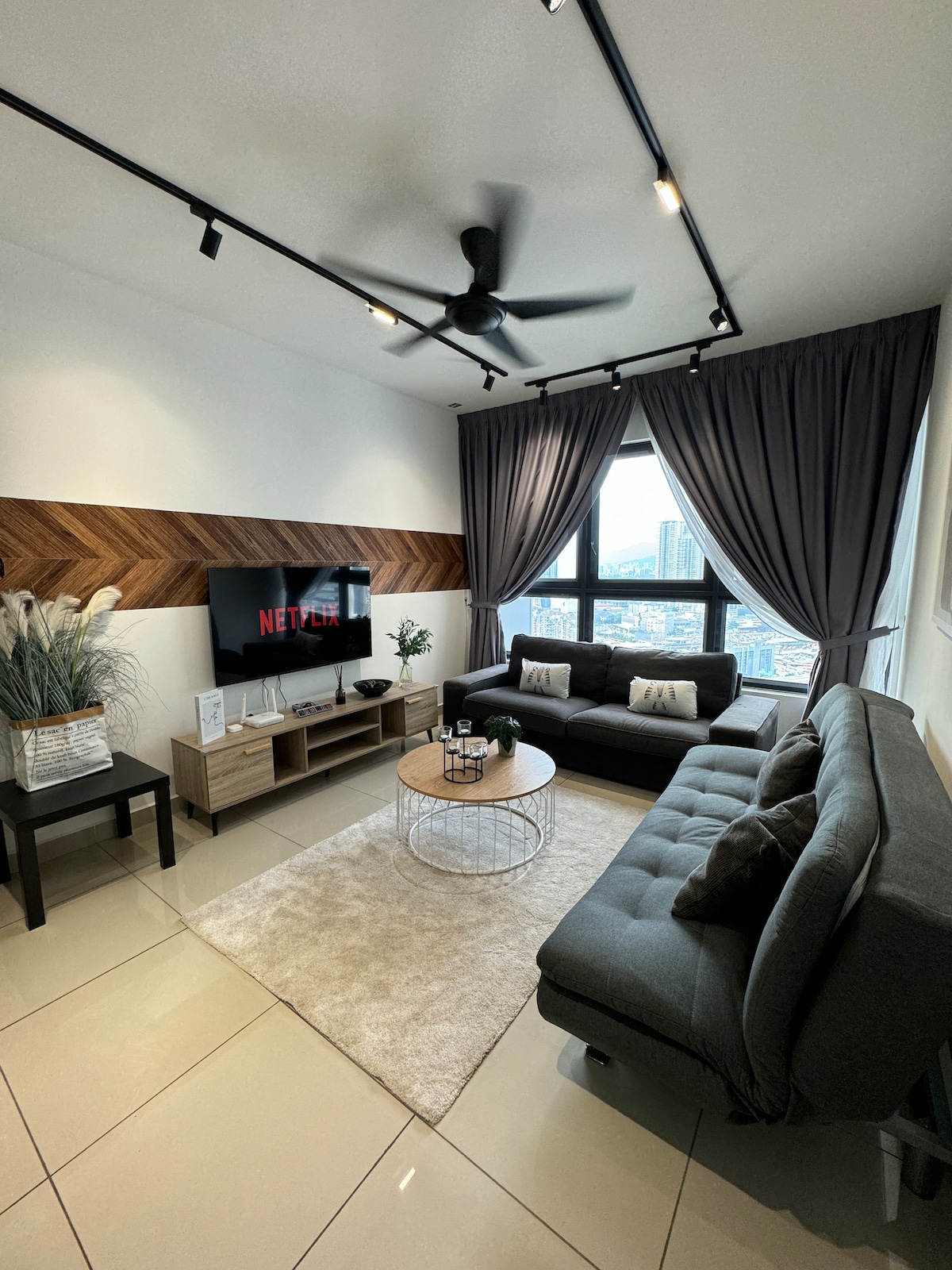 The living area features a modern design, showcasing a plush gray sofa, a matching futon, and a circular coffee table. A television mounted on the wall indicates entertainment options, while large windows allow natural light and provide views of the city skyline.