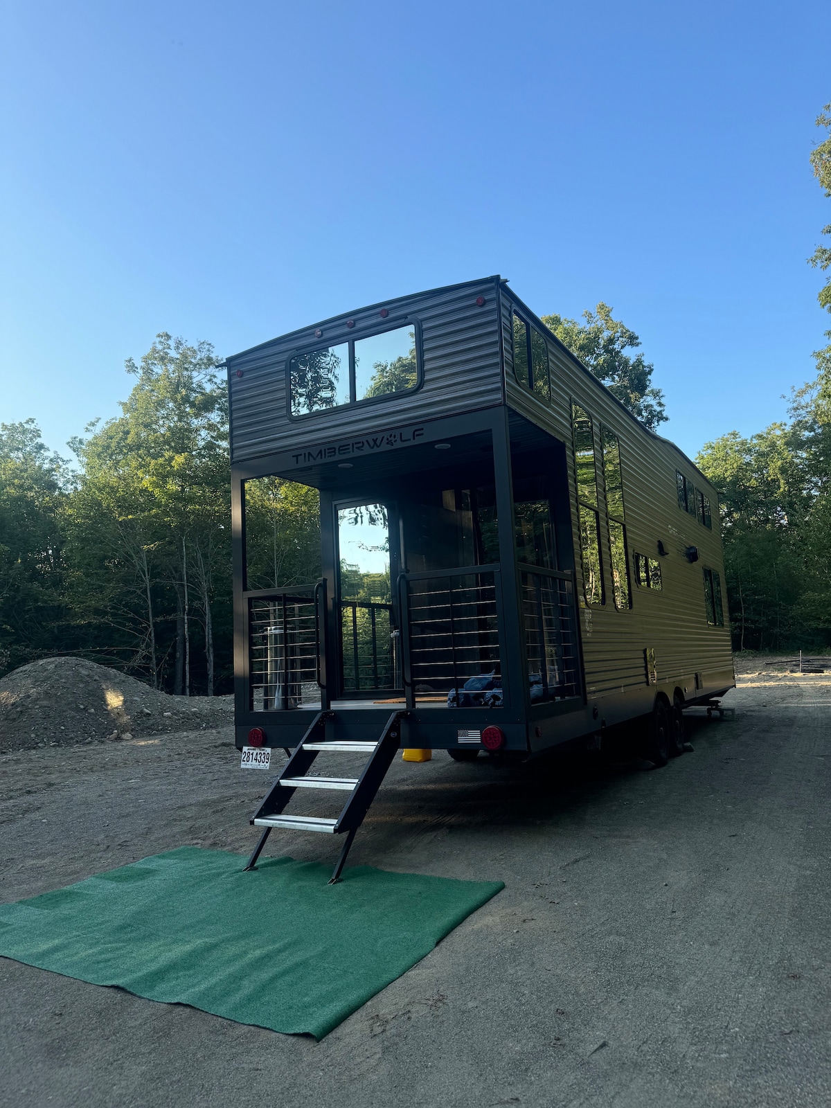 A tiny home on wheels is positioned outdoors, showcasing a modern exterior with large windows and a welcoming entrance. Steps lead up to the entryway, while a green mat is placed at the base. Surrounding trees provide a natural backdrop.