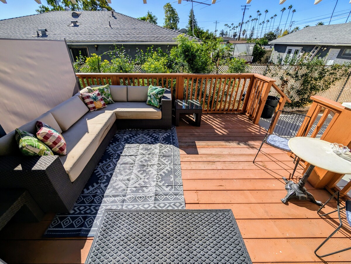 A spacious private sundeck is shown, featuring a comfortable seating area with a sectional sofa and decorative pillows. A small round table is present, alongside a rug that enhances the outdoor space. Lush greenery and nearby houses are visible in the background.