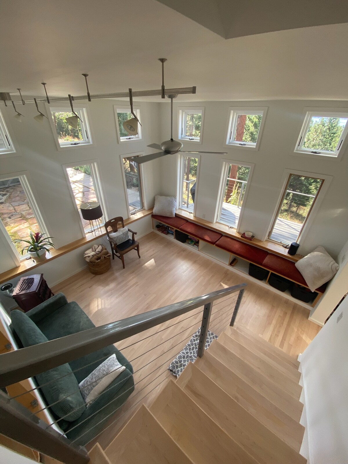 A bright living area features multiple windows allowing natural light to fill the space. A green sofa is positioned near a wooden bench with storage. A small wooden chair adds to the cozy ambiance, complemented by a ceiling fan and warm wooden flooring.