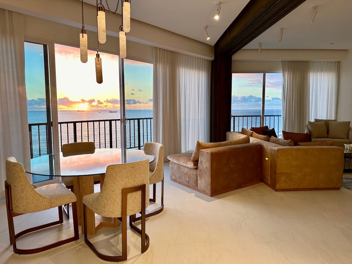 A modern living space features a glass dining table surrounded by four beige upholstered chairs. Large windows offer sweeping ocean views, and soft sunlight illuminates the room. A cozy seating area with a light-colored sofa is positioned near the windows, enhancing the inviting ambiance.
