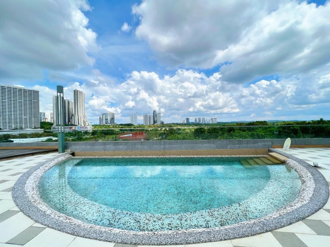An outdoor pool features a rounded shape bordered by a textured stone rim. The clear water reflects the sky and nearby city skyline, while a comfortable lounge chair is positioned adjacent to the pool for relaxation.