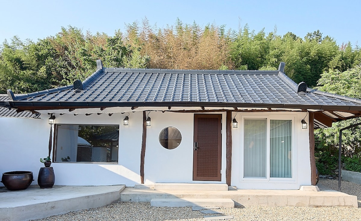 A traditional-style house is showcased with a sloped tiled roof and wooden accents. A round window and glass doors provide a view into the interior, while potted plants are positioned near the entrance. The surrounding landscape includes greenery, enhancing the tranquil setting.