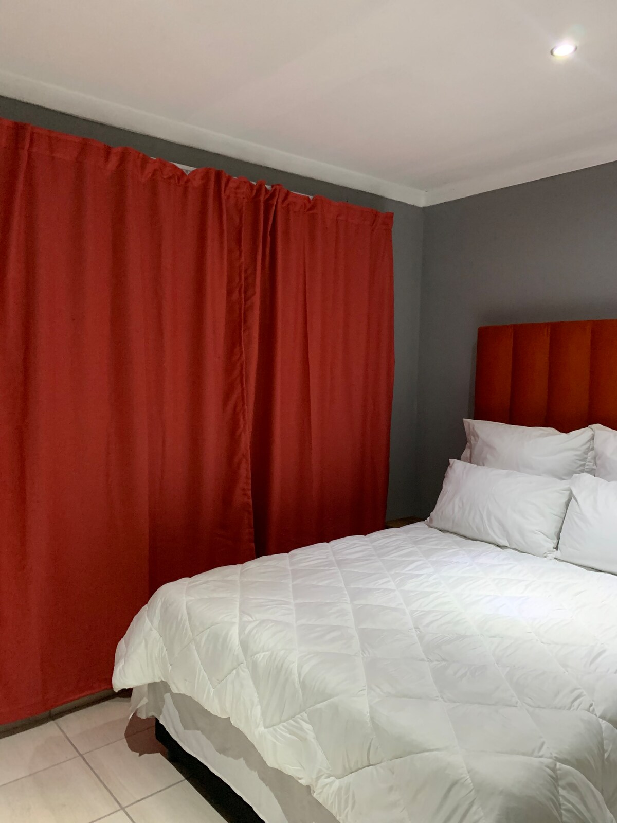 A cozy bedroom is presented with a spacious bed covered in a white quilt. Red curtains frame a wall, complementing the muted grey behind a headboard. Soft lighting overhead creates a calm ambiance in the room.