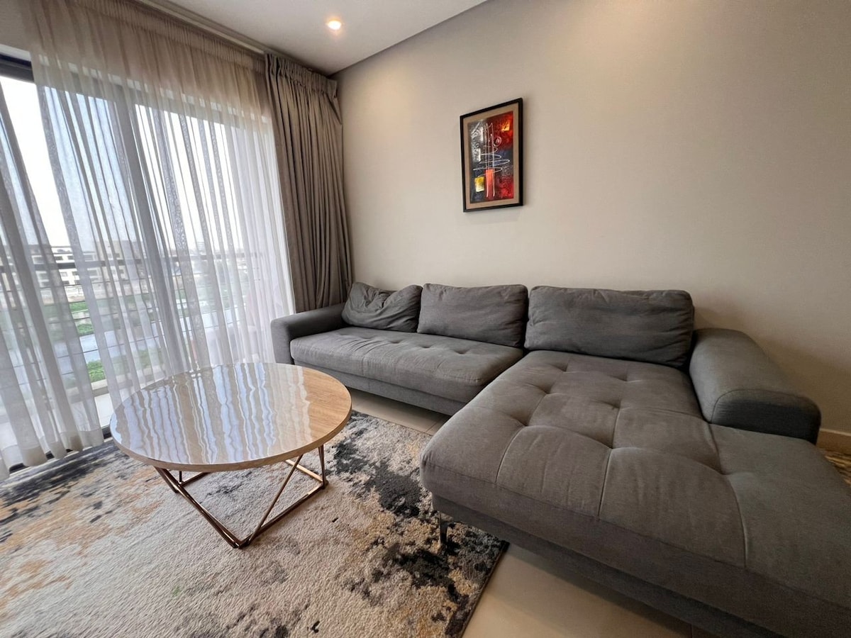 A modern living area features a plush gray sectional sofa, positioned next to a stylish round coffee table. Large windows are dressed with sheer curtains, allowing natural light to fill the space and offering a view of the outside.