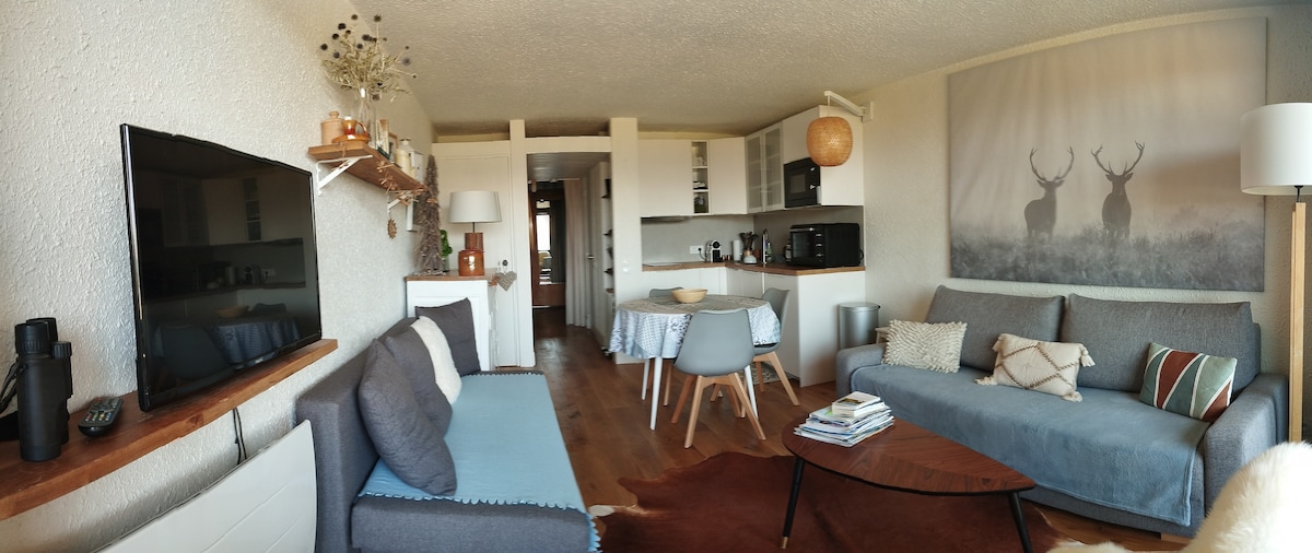 A cozy living space is presented with a blend of soft seating arrangements, including two sofas and a round dining table. Natural light fills the room, highlighting a modern kitchen area equipped with essential appliances. A large wall art piece featuring deer adds a focal point.
