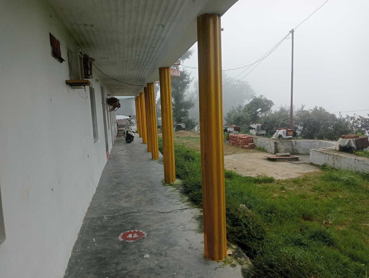 A covered walkway features golden columns supporting the roof, with misty surroundings enhancing the tranquil setting. The path leads to a grassy area, partially dotted with stone structures and greenery, providing a calm atmosphere for relaxation.