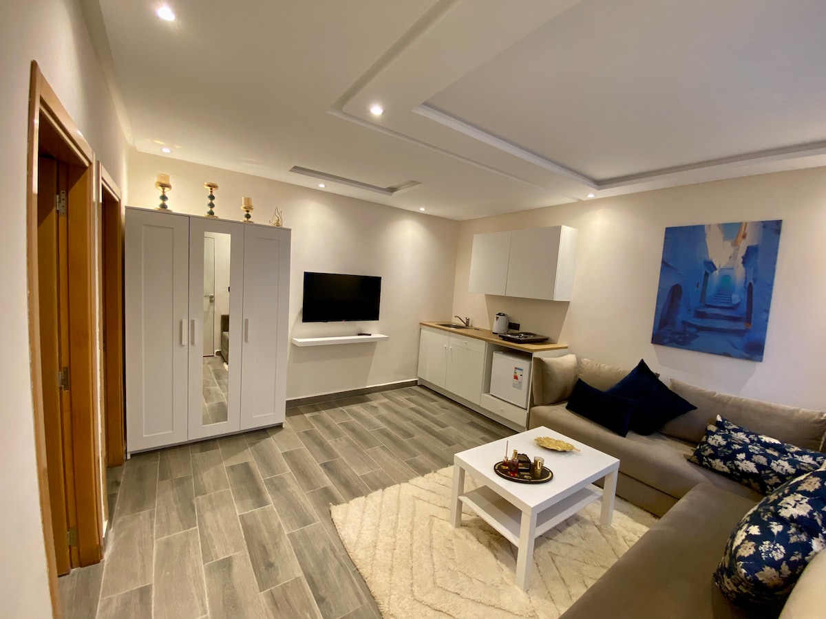 The living area features a beige sofa adorned with navy and white cushions. A minimalistic white coffee table sits on a textured rug. A wall-mounted television is positioned opposite a compact kitchen area, which includes white cabinetry. Natural light is provided by recessed lights and ceiling fixtures.