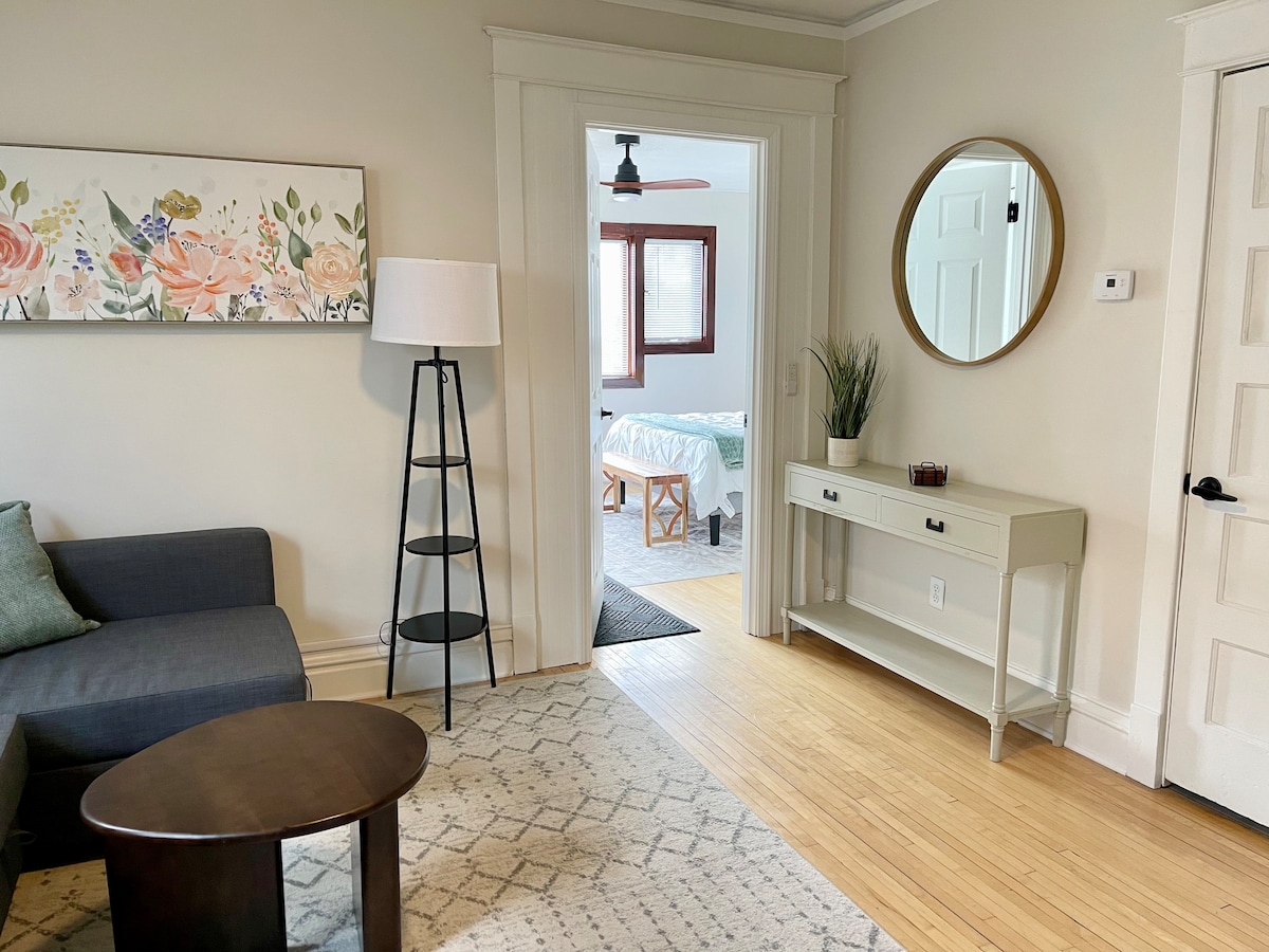 A welcoming entrance area shows a stylish standing lamp beside a dark gray sofa. A round mirror hangs above a small console table, complemented by a decorative plant. A floral artwork adorns the wall, leading towards a light-filled room through an open doorway.
