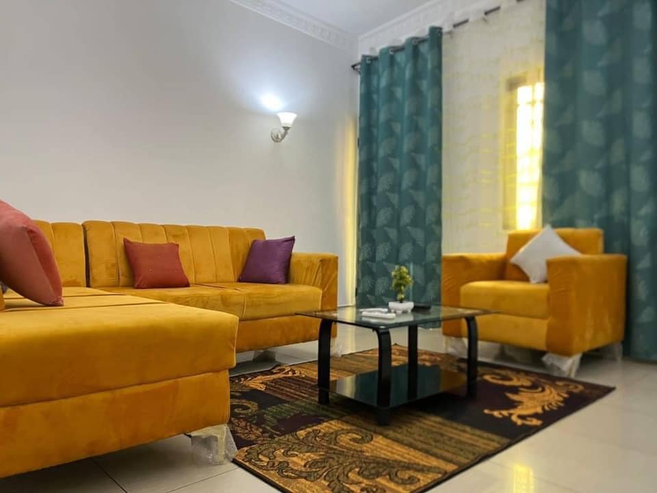 A bright living area features a yellow sectional sofa adorned with purple and white cushions. An accent chair complements the seating arrangement. A glass coffee table sits on a decorative rug, while sheer curtains allow soft light to filter in from the window.