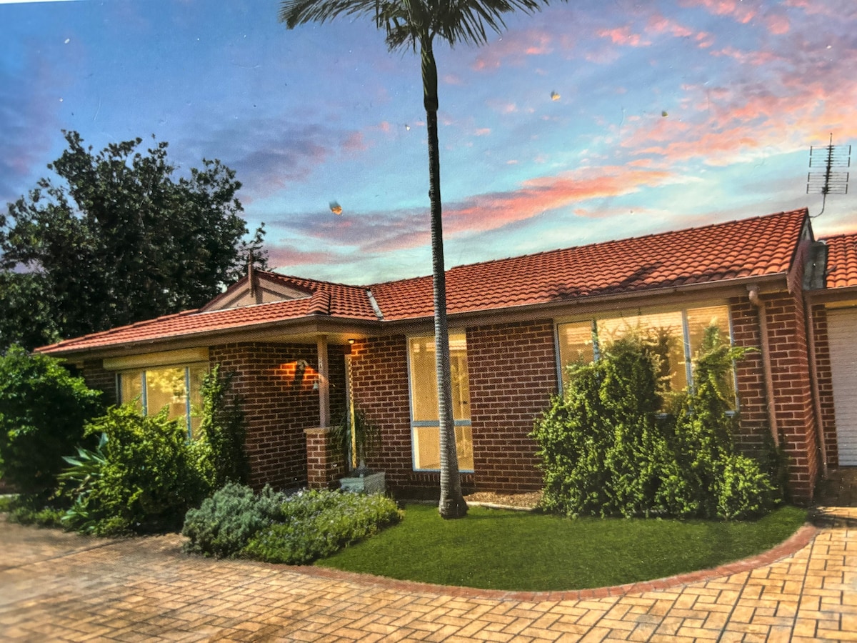 A brick house with a red tile roof is depicted under a colorful sunset sky. Lush greenery surrounds the entrance, with large windows allowing natural light inside. A palm tree adds tropical flair to the front yard, complemented by neat landscaping and a paved pathway.