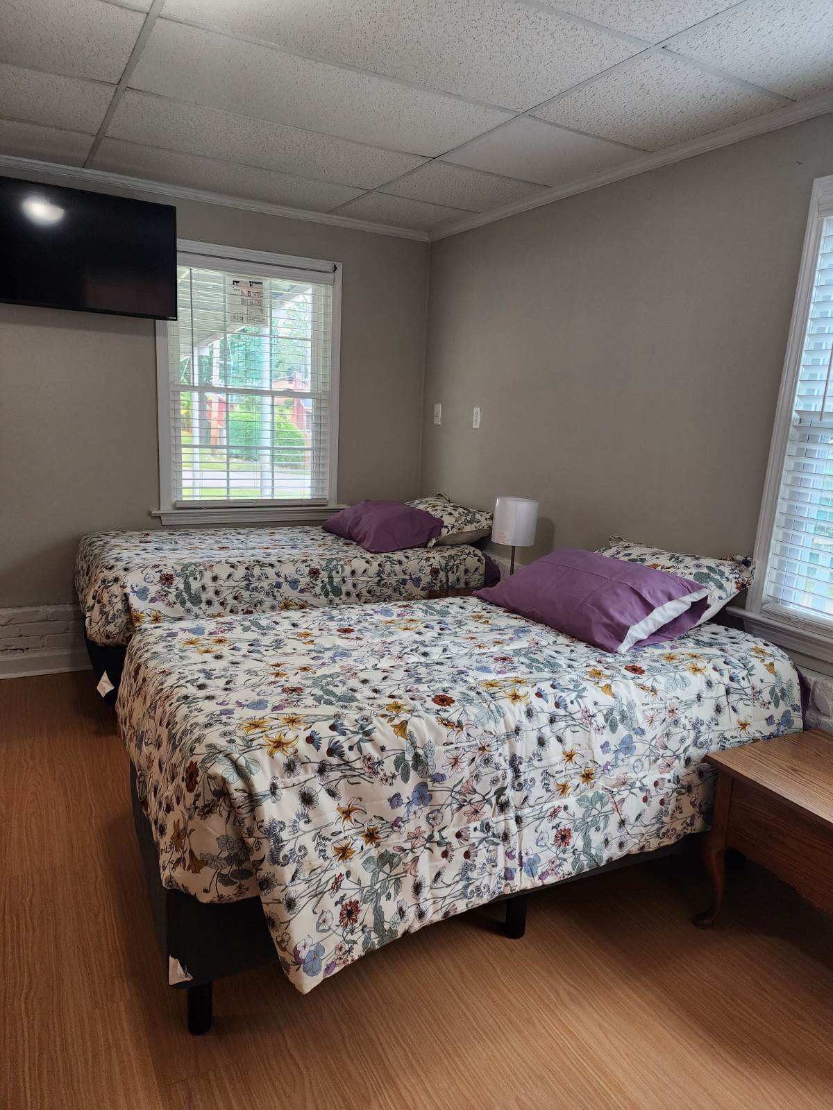 A well-lit room features two twin beds dressed with floral-patterned bedding. Each bed is complemented by purple accent pillows and positioned near a window with blinds. A small wooden bedside table is situated between the beds, and a wall-mounted TV is visible.