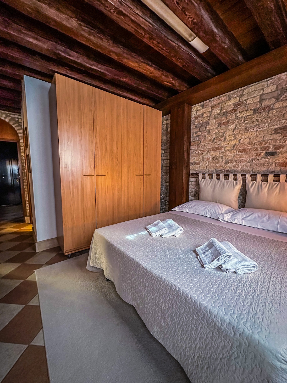 A spacious bedroom features a comfortable bed with light-colored bedding. Two neatly folded towels are arranged on the bed. A wooden wardrobe stands against the wall, and exposed stone walls complement the wooden beams of the ceiling, creating a cozy atmosphere.