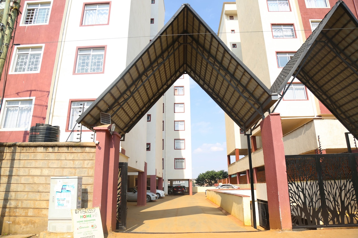 The image captures the entrance of the Greenzone apartment complex, framed by a distinctive archway. The structure consists of multiple floors with a combination of red and cream-colored walls. A gated access leads to an open parking area, lined by vehicles.