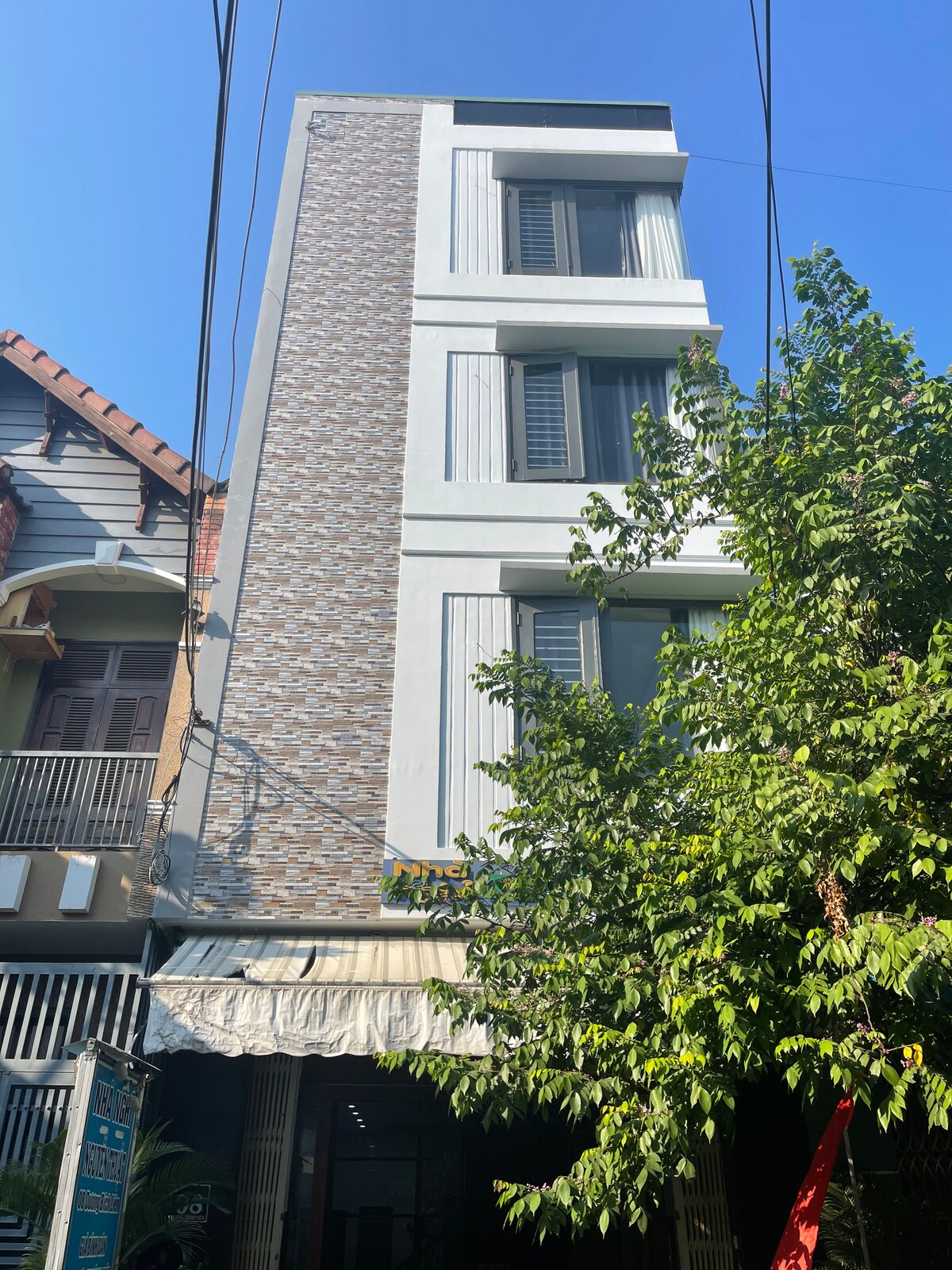 A modern three-story building is shown, featuring a mix of textured brick and smooth exterior walls. Large windows are positioned on each floor, allowing natural light to fill the interior. Green foliage is visible at the forefront, enhancing the urban landscape.