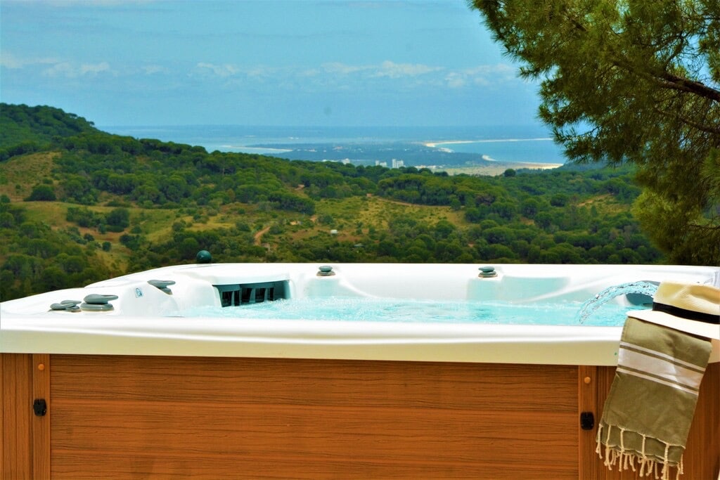 An outdoor jacuzzi accommodates up to six people, surrounded by scenic views of rolling green hills and distant coastline. Gentle steam rises from the water, inviting relaxation as a striped towel rests on the edge, harmonizing with the natural landscape.