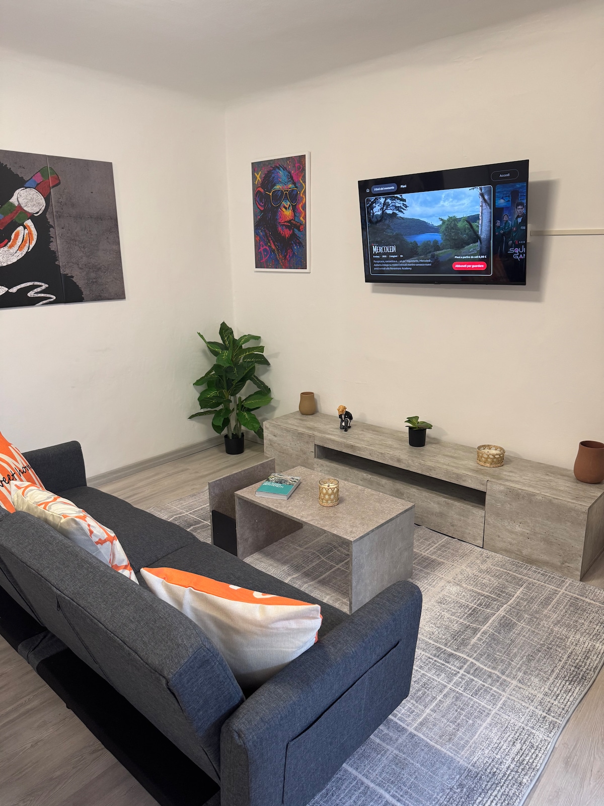 A cozy living area is presented with a spacious gray sofa adorned with colorful cushions. A minimalist coffee table sits in front, accompanied by decorative items. A flat-screen television is mounted on the wall, while a plant adds a touch of greenery to the space.
