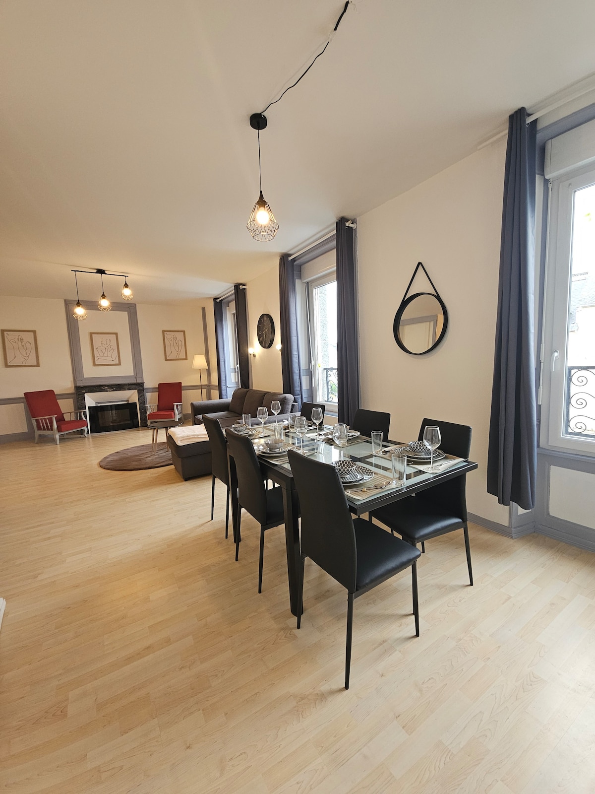 A dining area is showcased with a glass-top table set for six, surrounded by sleek black chairs. Natural light enters through large windows adorned with sheer curtains. A cozy seating area is visible in the background, featuring two red armchairs and a stylish mirror accent.