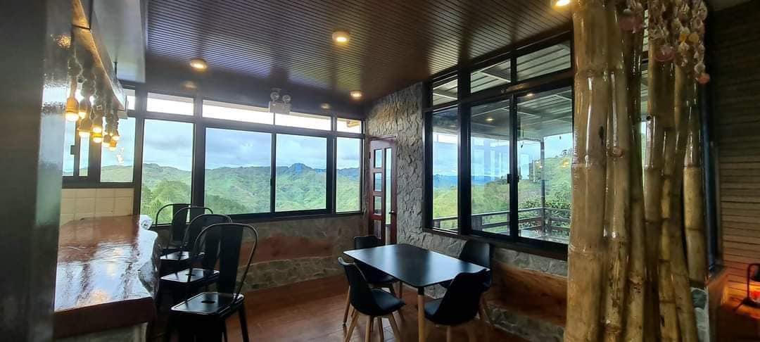 A spacious dining area features large windows that offer sweeping views of the surrounding landscape. Natural light fills the room, highlighting a black table surrounded by chairs. A bar counter made of stone is visible in the background, creating a comfortable atmosphere.