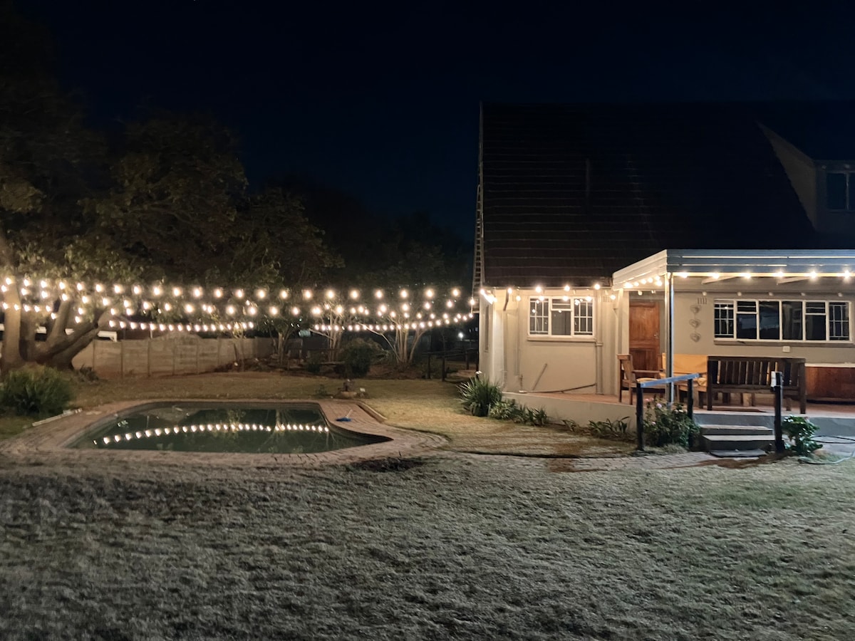 A well-lit outdoor area is showcased at night, with warm string lights illuminating the surrounding trees. A swimming pool is visible in the foreground, with a wooden seating area connected to the house, providing a welcoming space for relaxation.