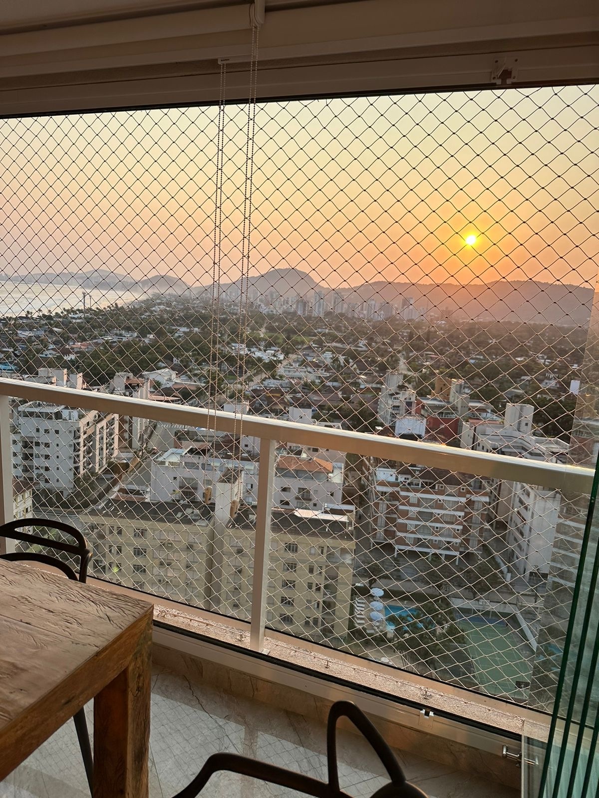 A sunset view is presented from a glass balcony, revealing a sprawling cityscape below. The horizon is illuminated in soft orange hues, while distant mountains are faintly visible. A protective netting is seen in front, ensuring safety while enjoying the picturesque scenery.