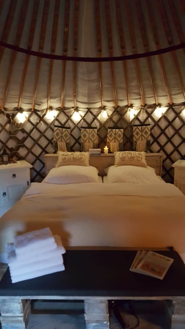 A cozy interior of the yurt features a spacious bed with white linens. Pendant lights softly illuminate the wooden walls, adorned with decorative elements. Two bedside tables are positioned on either side of the bed, enhancing the inviting atmosphere of the space.