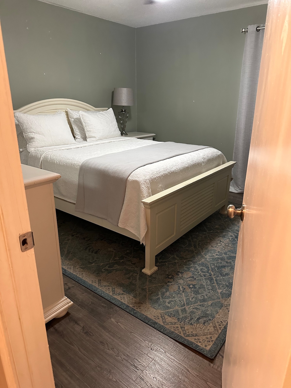 A private bedroom is shown, featuring a spacious bed with white linens and decorative pillows. A soft gray lamp provides ambient lighting on a bedside table. A light blue rug is placed on the dark wood floor. Soft gray curtains frame a window, enhancing the serene environment.