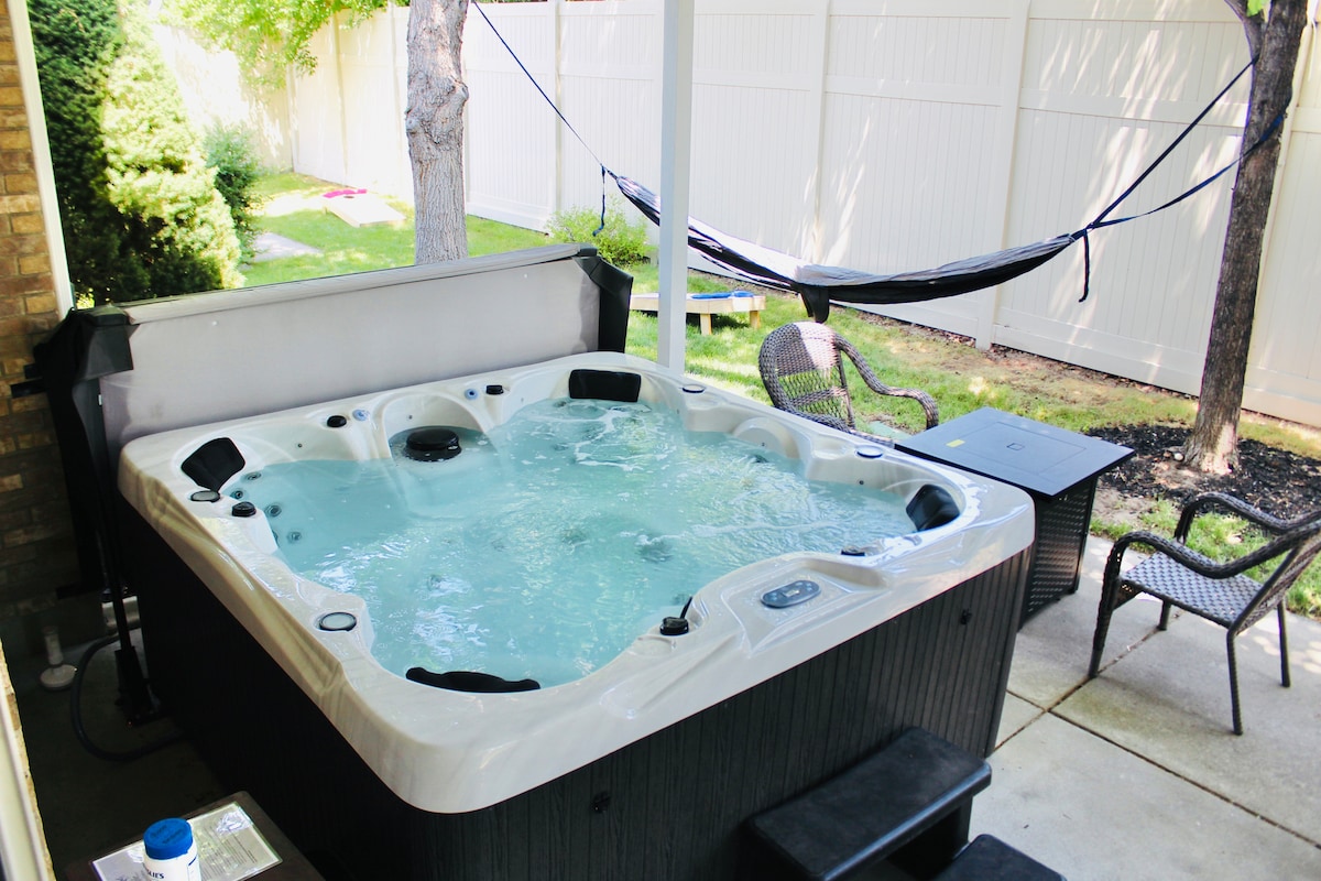 A spacious outdoor area features a private hot tub surrounded by a shaded patio. Nearby, a hammock hangs between two trees, inviting relaxation. Two wicker chairs and a small table are positioned beside the hot tub, adding to the comfortable outdoor setting.
