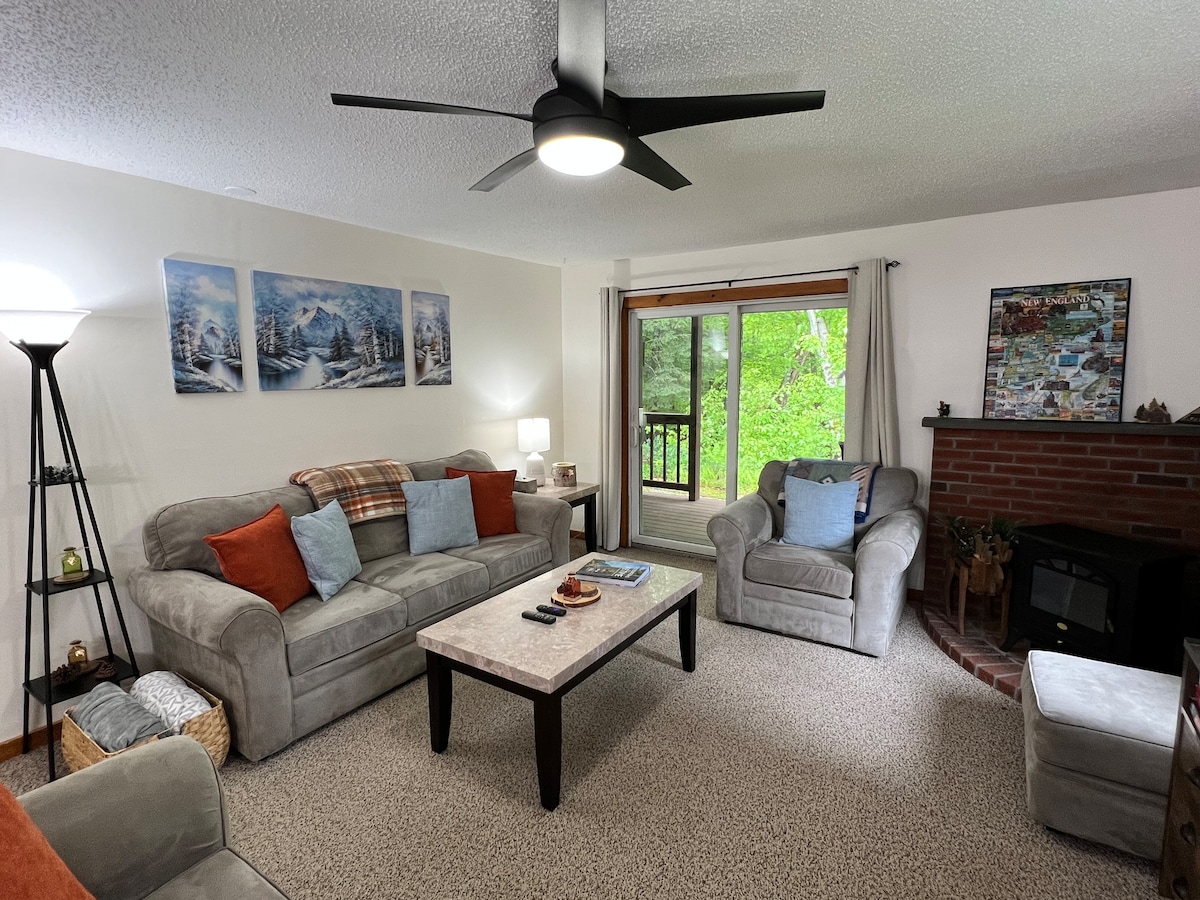 A cozy living room features two sofas with accent pillows, a coffee table, and a light fixture. Natural light enters through sliding glass doors, revealing a view of greenery outside. A brick fireplace and wall-mounted decor add character to the space.
