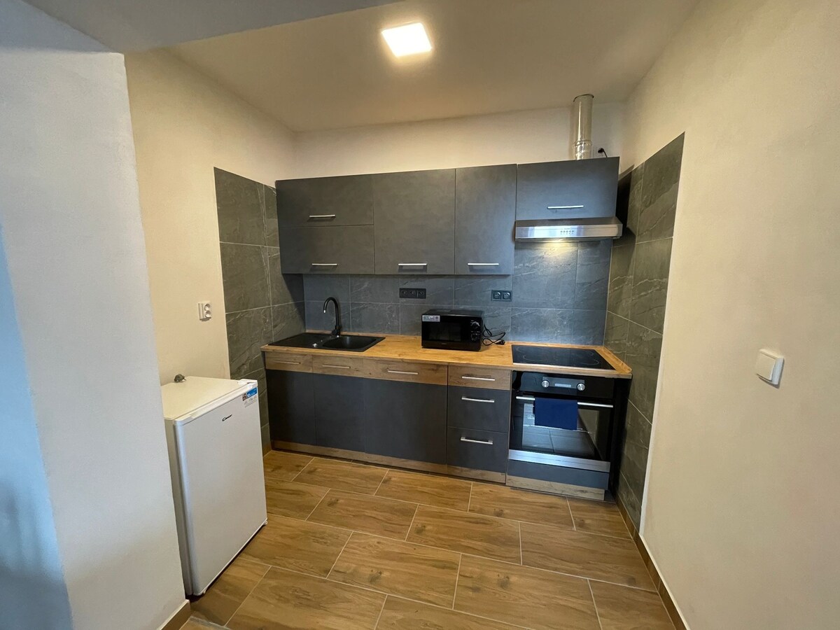 A modern kitchen is equipped with dark cabinetry and light wood countertops. A stainless steel stove and hood are paired with a microwave. A compact refrigerator is positioned on the left, while ample natural light highlights the tiled flooring.