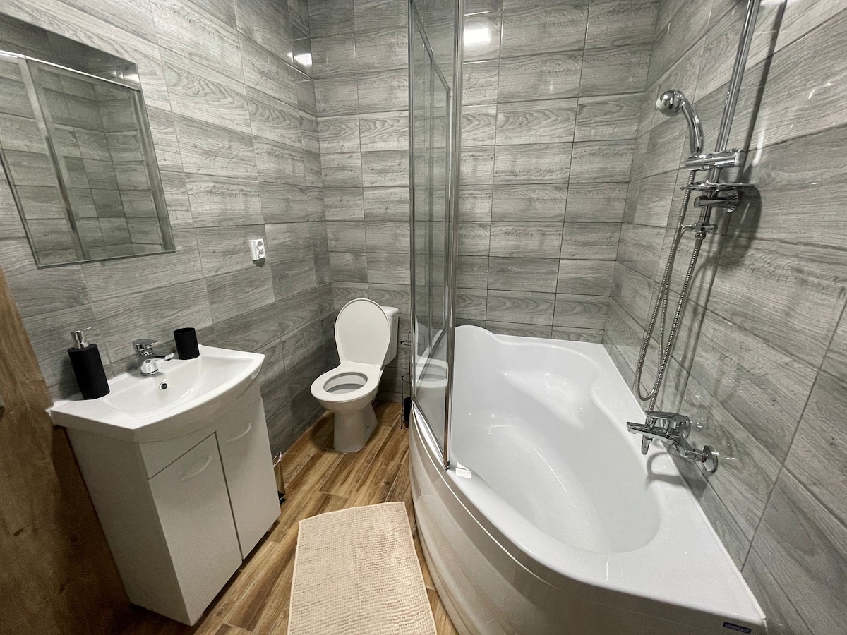 A modern bathroom is showcased with a spacious bathtub and a glass shower enclosure. A white vanity with a sink is situated next to a clean toilet. Neutral tiles adorn the walls, complemented by a warm wooden floor and a soft bath mat on the ground.