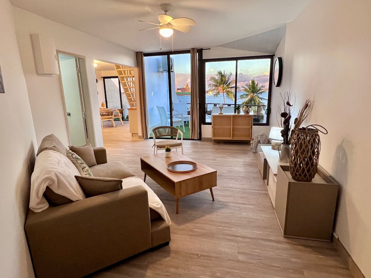 A spacious living area features a comfortable sofa and modern wood coffee table. Large windows provide expansive views of the ocean and surrounding palm trees. Natural light fills the space, creating an inviting atmosphere. Minimalist decor showcases clean lines and neutral tones.