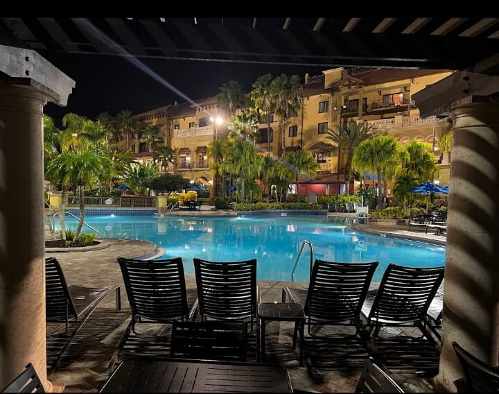 A serene swimming pool is illuminated by night, surrounded by lush palm trees and tropical landscaping. Comfortable lounge chairs are arranged neatly at the water's edge, offering a relaxing spot. Multiple buildings are visible in the background, showcasing a welcoming resort atmosphere.