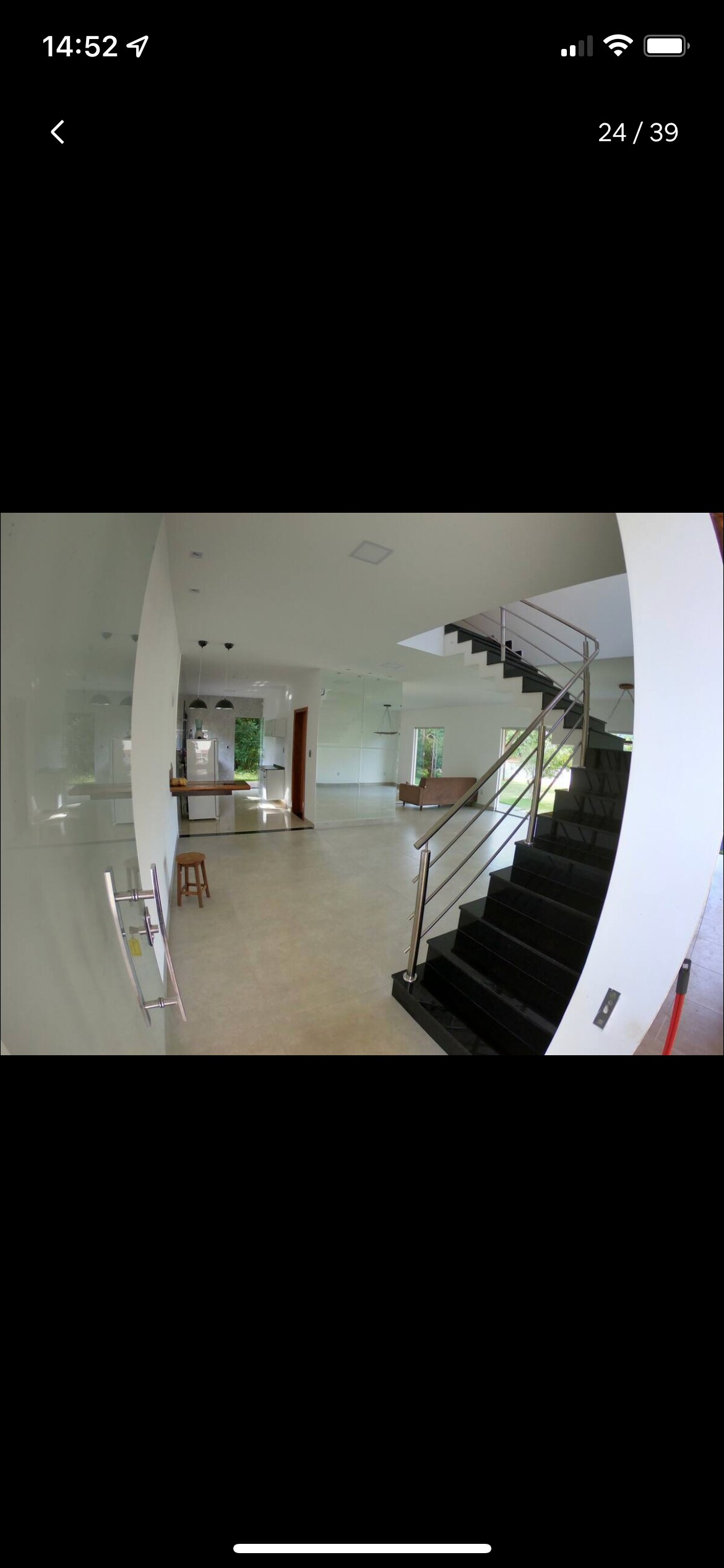 An open and airy living space is presented, featuring clean white walls and a contemporary staircase with a sleek black finish. Natural light floods the area, highlighting the polished floor. A glimpse of the kitchen area is visible in the background, enhancing the sense of depth.