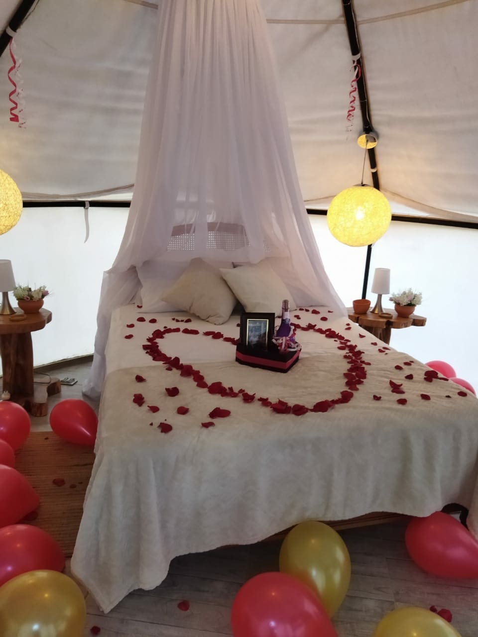 A cozy tented space features a neatly made bed draped in a light canopy. The bed is adorned with rose petals arranged in a heart shape, surrounded by balloons in red and gold. Soft lighting is provided by two round lamps on wooden side tables.