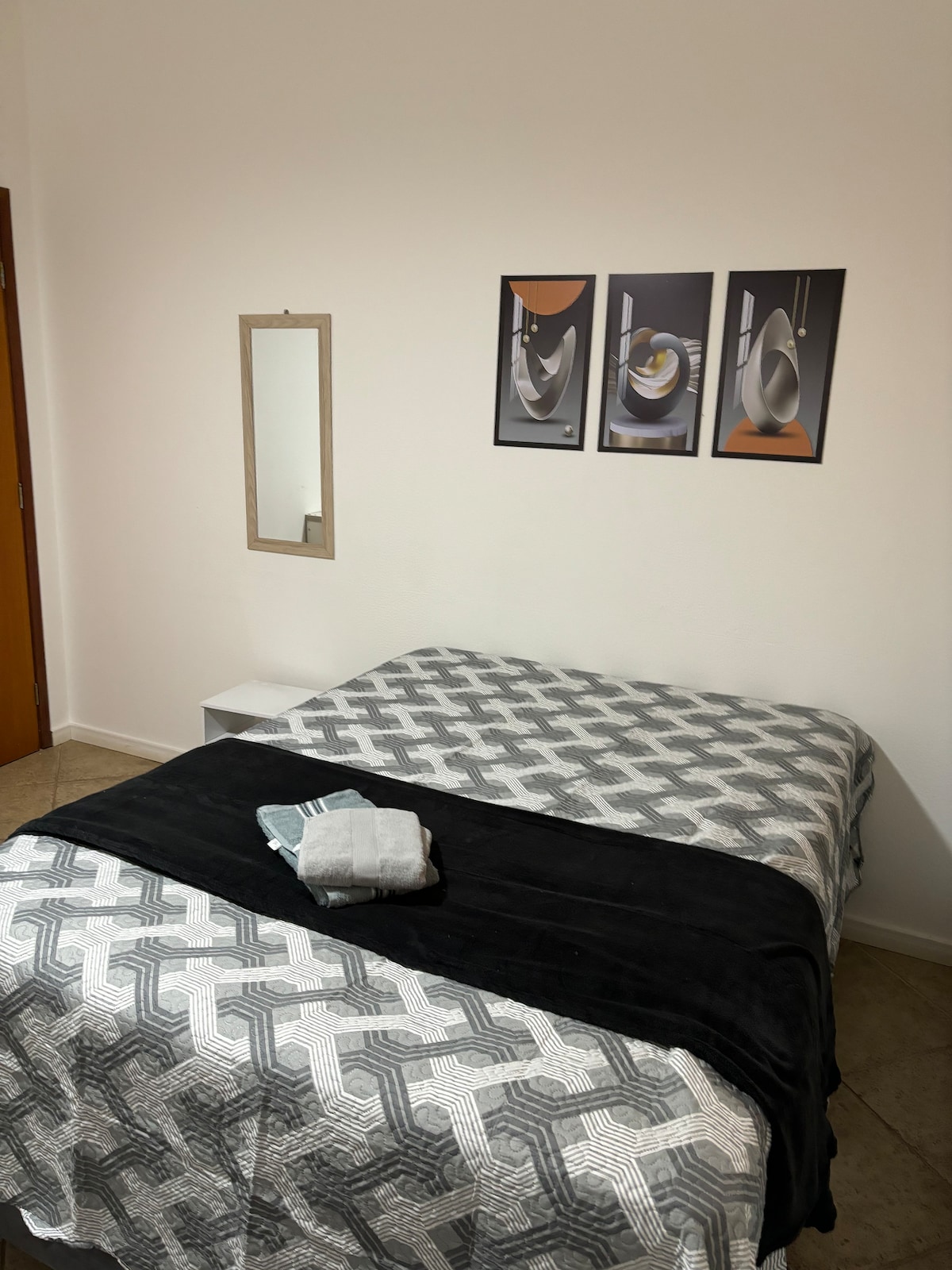 A spacious sleeping area features a geometric patterned bedspread draped over the bed. A soft black throw is neatly placed at the foot of the bed. Three abstract framed artworks adorn the wall, and a mirror is mounted alongside a bedside table.