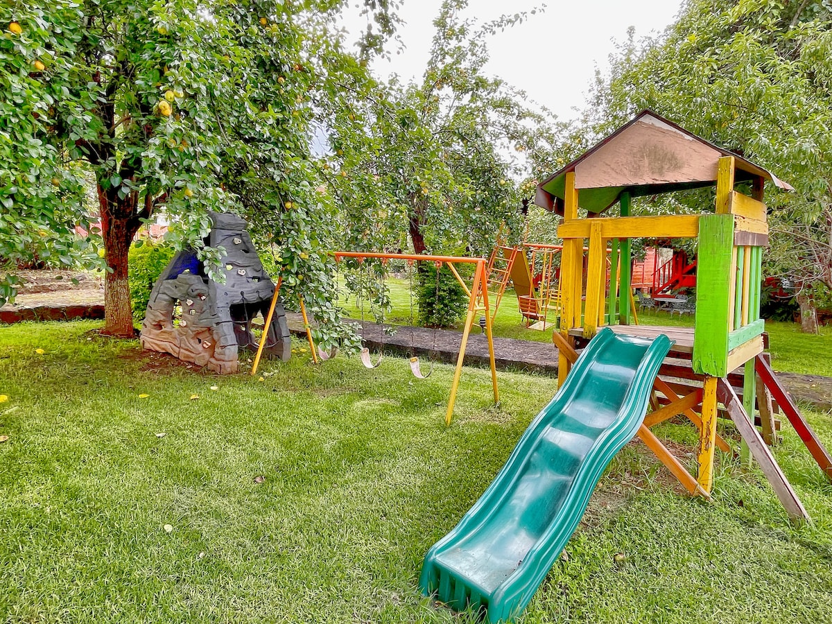 A colorful playground area is visible, featuring a green slide, swings, and a playhouse surrounded by leafy trees. The grass is well-kept and provides a safe space for children to play and explore.