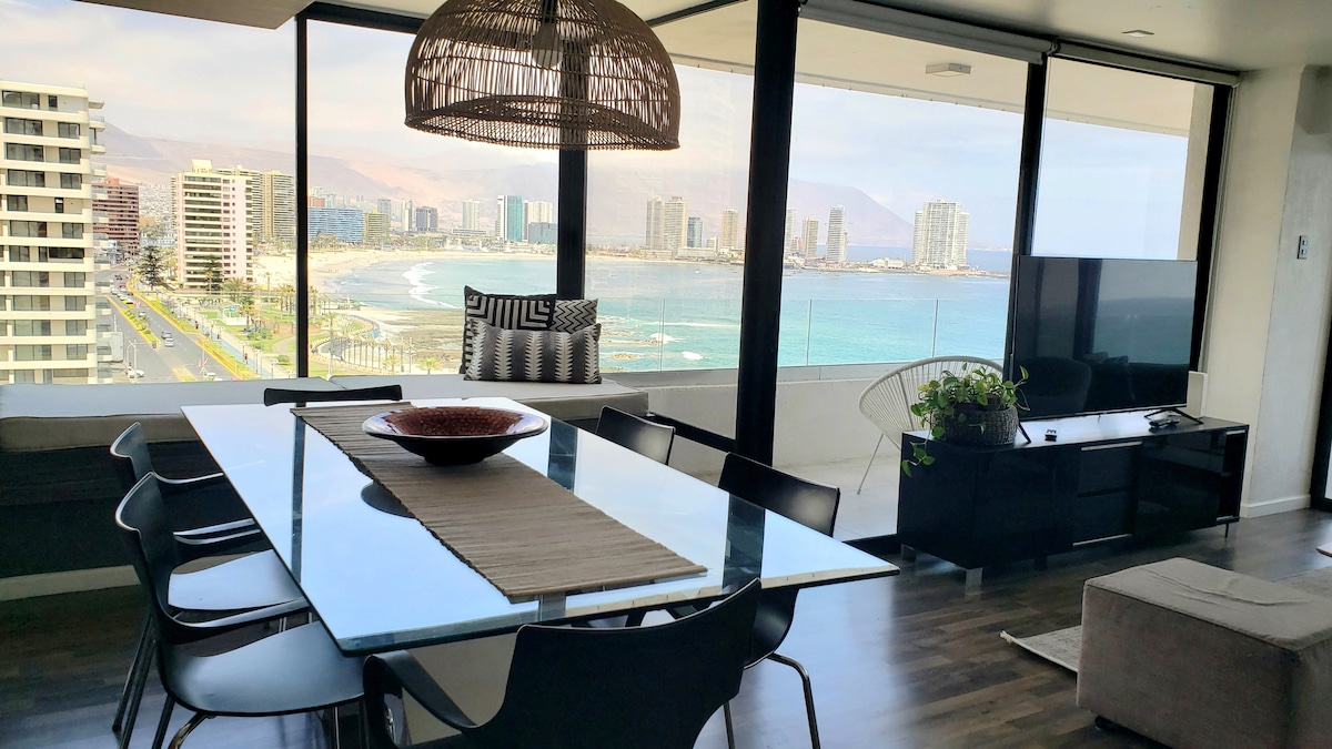 A spacious dining area features a glass table surrounded by black chairs, with a large decorative bowl at the center. Expansive windows showcase views of Iquique's coastline, blending indoor dining with picturesque outdoor scenery.