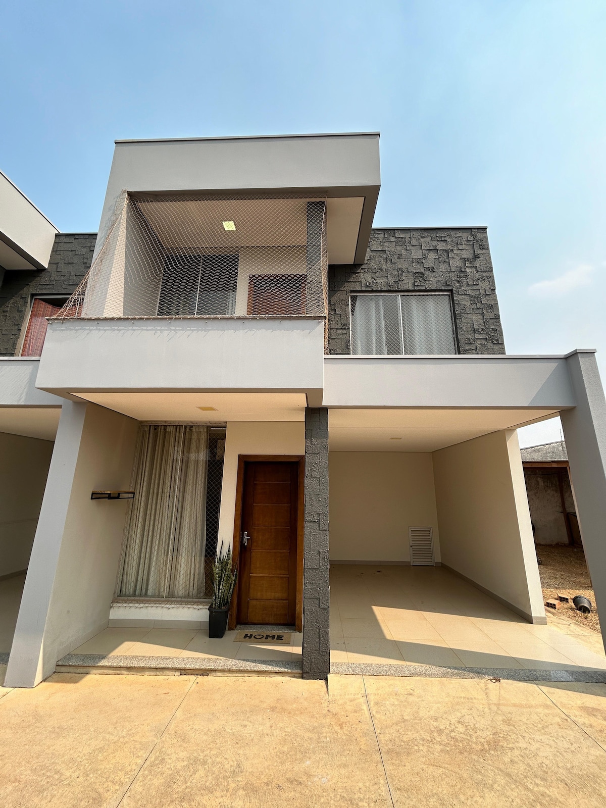 The exterior of the apartment features a modern façade with a combination of light and dark stone materials. A wooden door leads into the space, complemented by a large window covered with a sheer curtain. A small potted plant is placed next to the entrance.