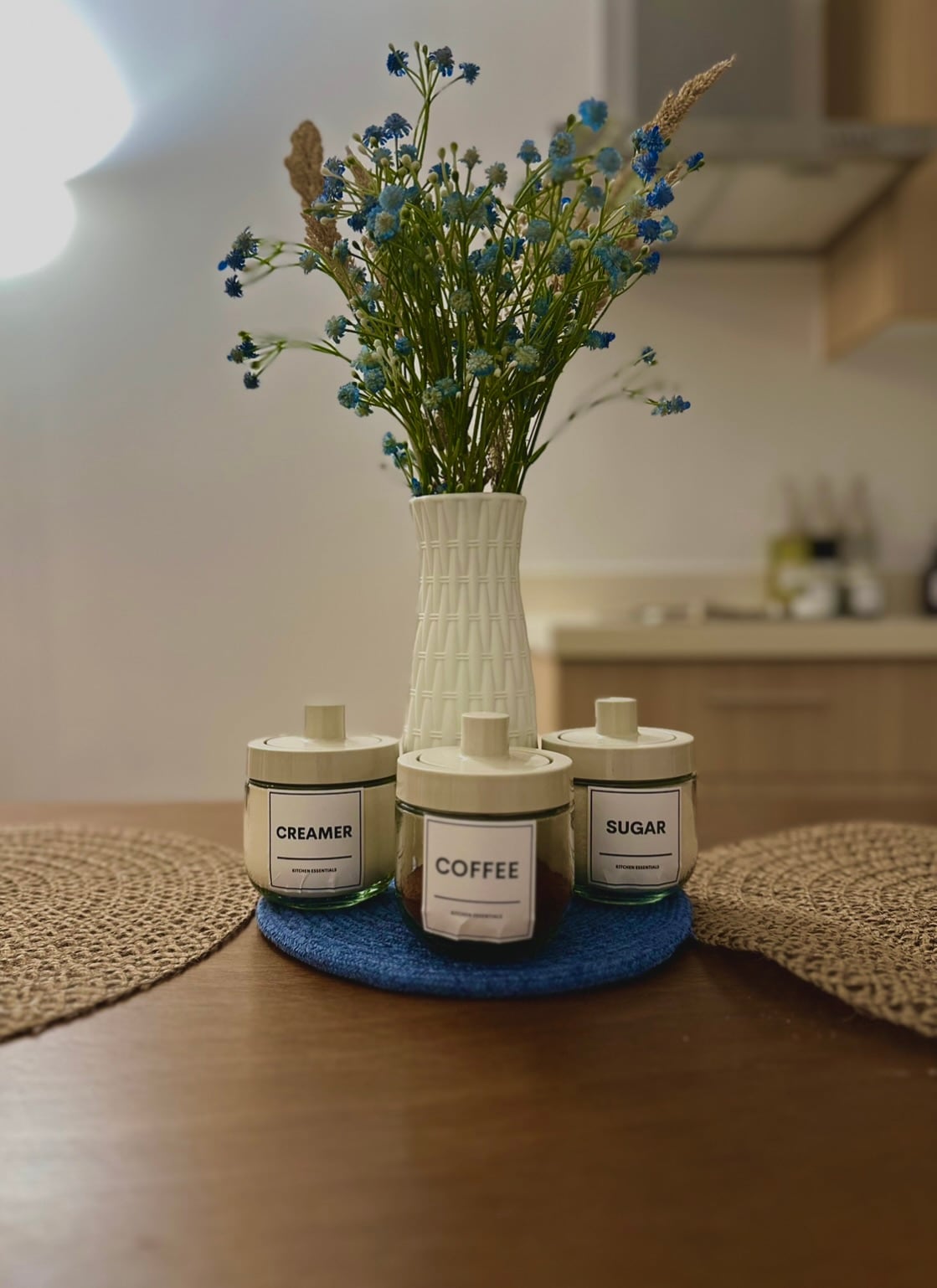 A minimalistic setup features a white vase filled with blue flowers at the center of a wooden dining table. Three labeled containers for coffee, creamer, and sugar are arranged on a circular blue mat, providing an organized and functional touch.