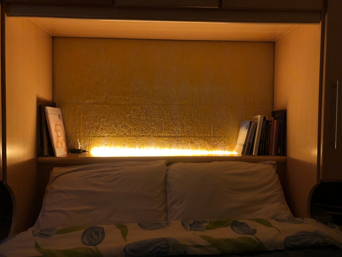 A cozy sleeping area features a wooden headboard with built-in lighting that softly illuminates the space. The bedding is complemented by a decorative throw, while books are neatly arranged on either side of the headboard.