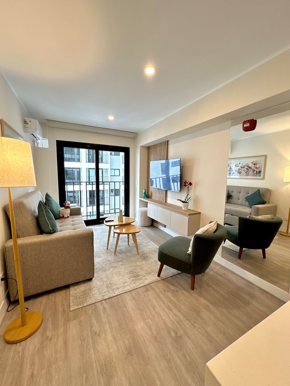 A bright living area features a comfortable gray sofa, a small wooden coffee table, and two accent chairs. Large glass doors provide access to a balcony, while elegant decor and a light area rug enhance the inviting atmosphere. A mirror reflects the light and space.