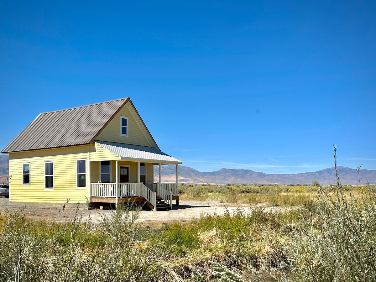 A charming yellow cottage with a gabled roof is set against a backdrop of rolling hills and clear blue skies. The porch features a railing, offering a welcoming entry. Surrounding vegetation consists of sparse desert flora, highlighting the remote, tranquil setting.